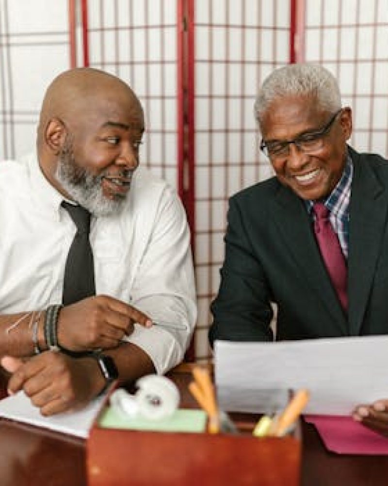 Two senior men in business attire discussing documents with a positive interaction in an office setting.